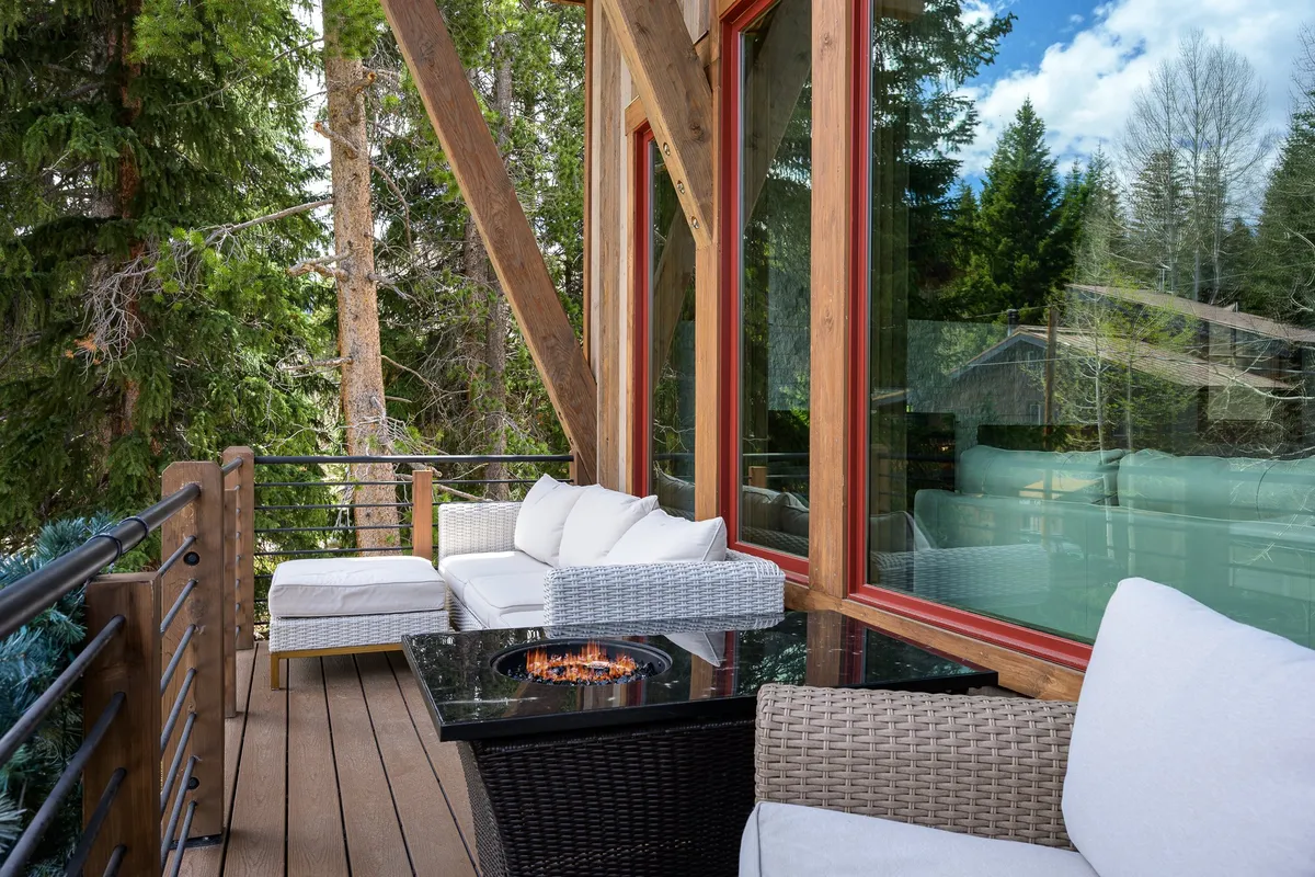 A cozy outdoor setting with a fire pit, comfortable seating, large chalet windows, and surrounding trees. The scene is tranquil, inviting relaxation after a day of adventure, and suggests a seamless indoor-outdoor living experience.