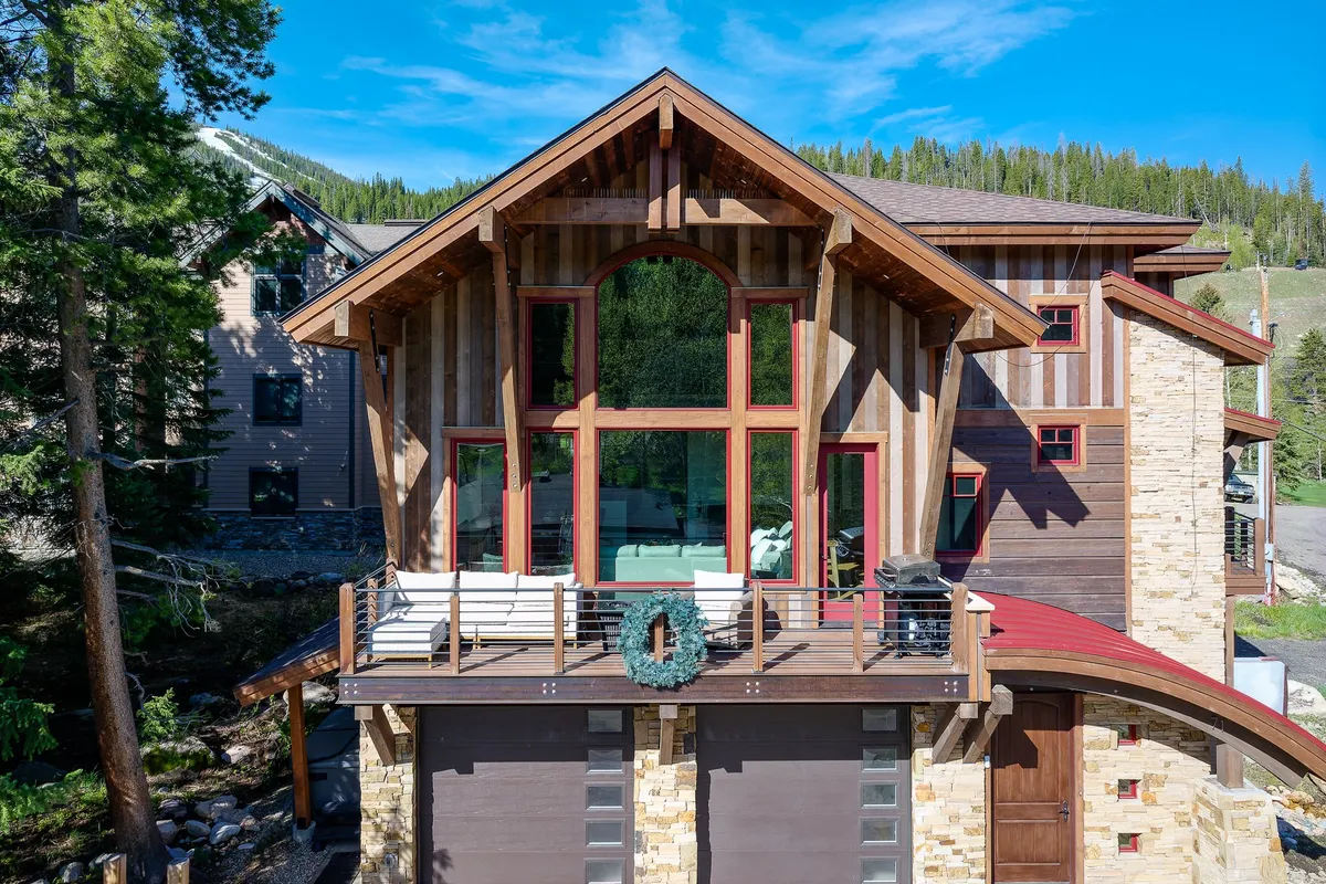 This exterior image displays the front of the home, featuring large windows, wooden accents, a stone facade, and a spacious deck with inviting seating. The scene is tranquil, with the natural landscape visible around the home, suggesting privacy and a seamless connection to the surrounding mountain environment.