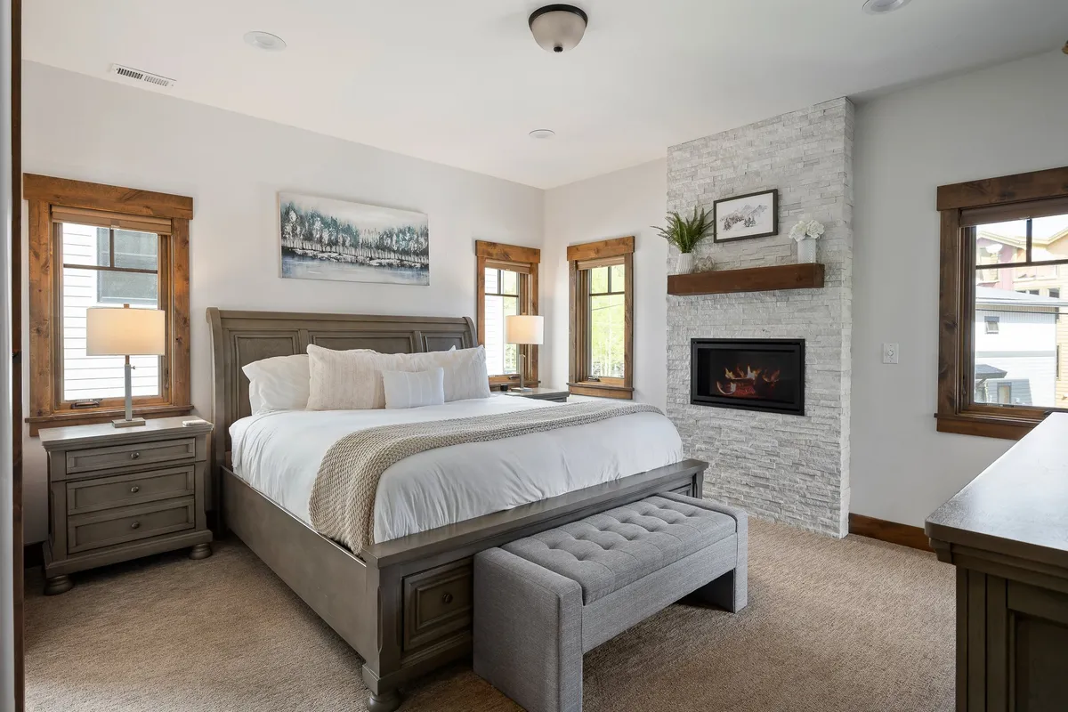 A spacious bedroom featuring a large bed with plush bedding, a cozy fireplace, decorative art on the wall, and nightstands with lamps. The space exudes tranquility and comfort, enhanced by soft neutral tones and thoughtful décor.