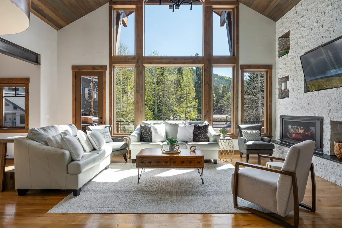This image displays a spacious, sunlit living room with large windows offering views of the outdoors. The seating area is arranged around a cozy fireplace, with a decorative coffee table in the center and elegant mountain-modern décor throughout.