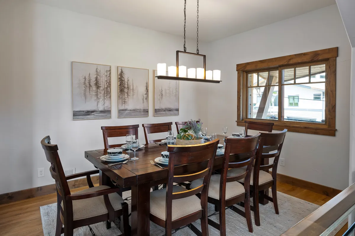 A stylish dining area highlighted by a large dining table, an elegant chandelier, artwork on the wall, and a bright window letting in natural light. The table is arranged for family-style meals and celebrations.