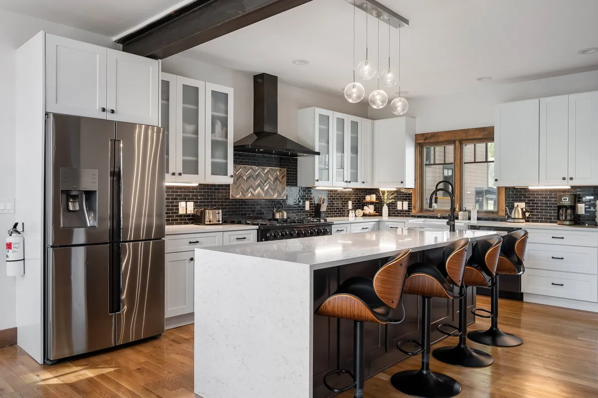A modern kitchen featuring a large kitchen island as the focal point, with a stainless steel refrigerator, striking range hood, pendant lighting, and a herringbone backsplash. The space is bright and designed for both cooking and entertaining.
