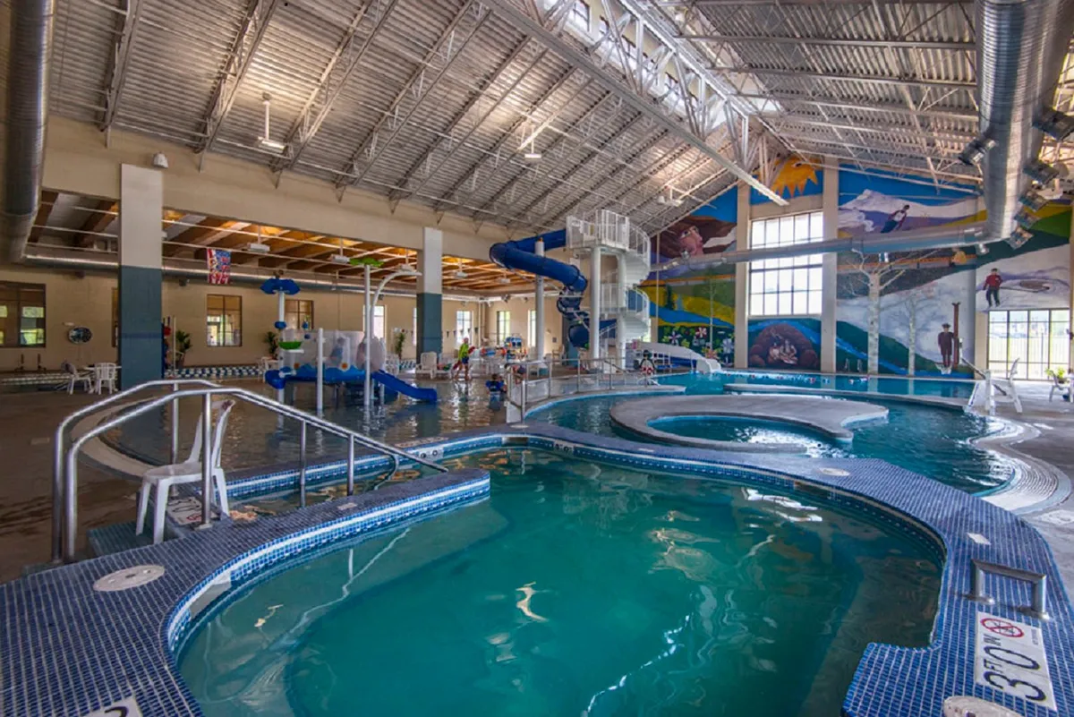 A vibrant interior scene inside a recreation center, filled with children laughing and playing in the swimming pool and racing down a waterslide. The lively mural and energy-packed atmosphere emphasize fun, movement, and family togetherness.