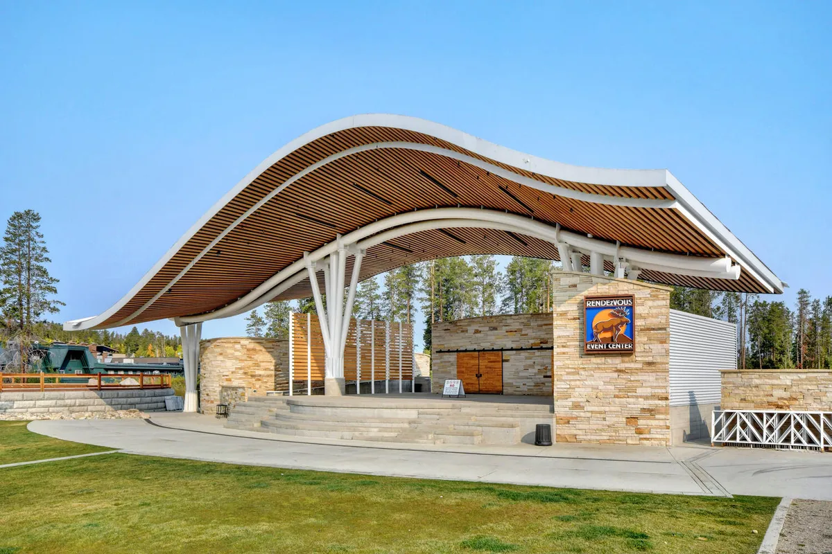 This image shows the exterior of a community event center, featuring a distinctive curved roof, stone facade, and a wooden seating area. The event center signage is visible, and the open space suggests a venue for gatherings, concerts, or festivals.