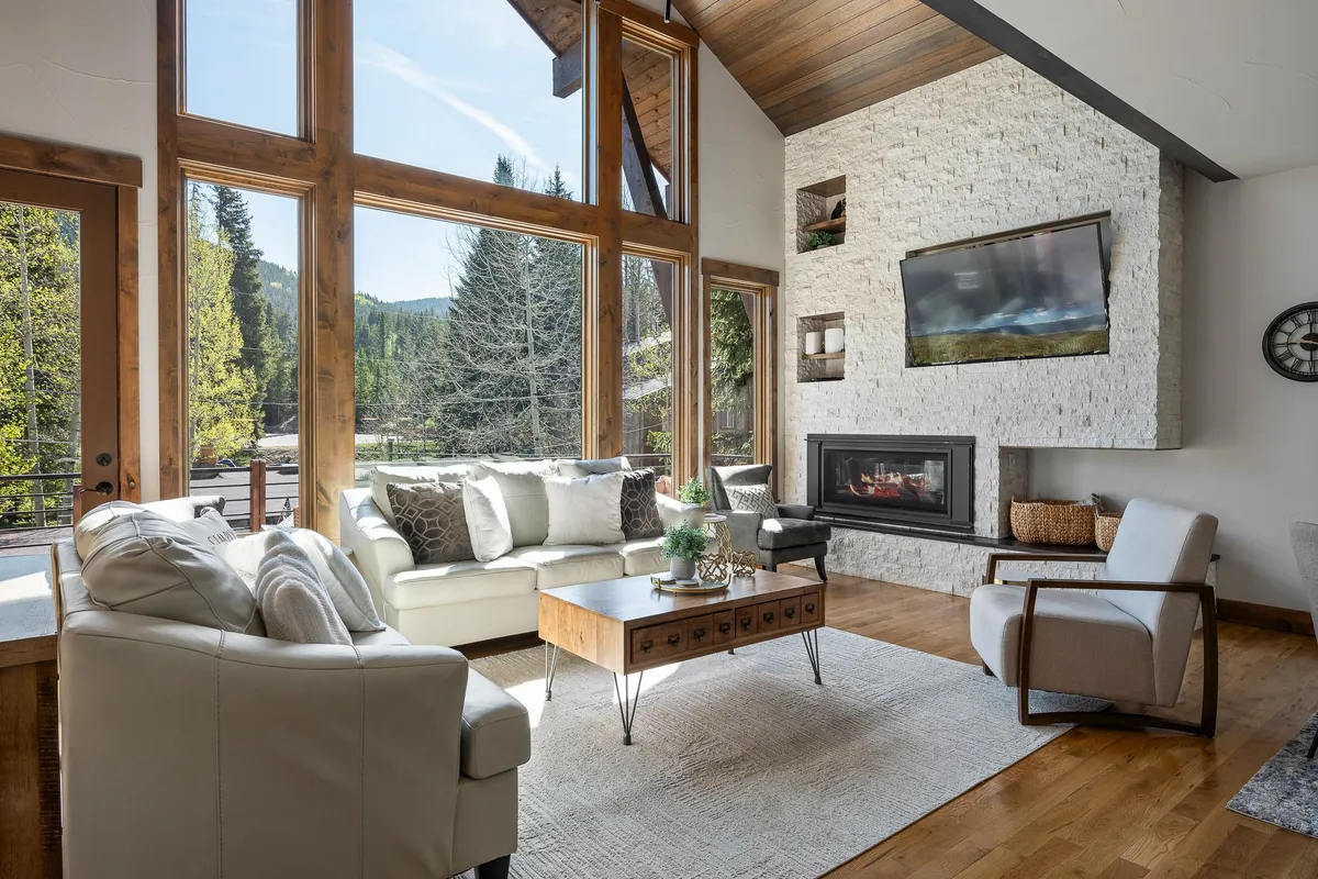 A luxurious interior living space with large windows framing dramatic mountain views, a grand stone fireplace, and plush, comfortable seating. The room exudes a serene, welcoming atmosphere—ideal for relaxing after outdoor adventures while appreciating the natural beauty beyond.