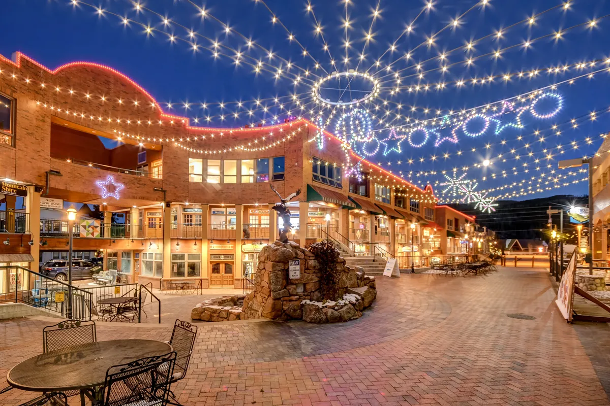 Capturing a lively streetscape, this image features string lights overhead, decorative buildings, a central stone feature, and people engaging in dining, shopping, and socializing. The festive ambiance suggests both daytime and evening activities in a welcoming downtown environment.
