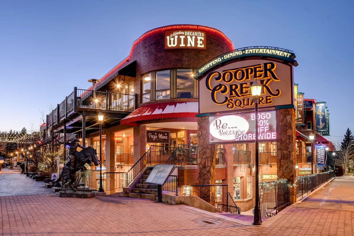 An inviting exterior shot of Cooper Creek Square, featuring its sign, vibrant storefronts, outdoor seating, and a contemporary sculpture. The scene is lively, suggesting opportunities for shopping, dining, and entertainment just minutes from the chalet.