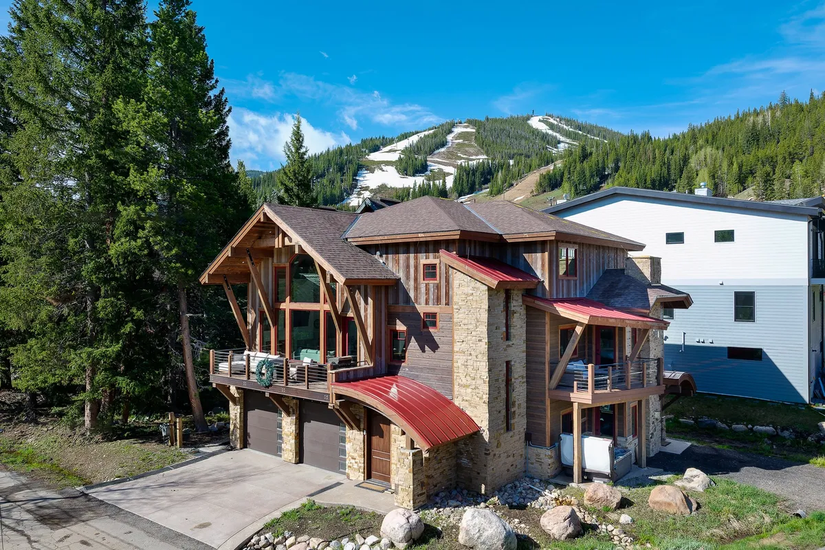 The image showcases a stunning custom chalet exterior with bold wooden beams, a striking stone facade, a vibrant red metal roof, and expansive windows. The home is nestled at the base of majestic mountains, with a spacious balcony for relaxing and fire tables perfect for gathering. The scene hints at year-round adventures like skiing, hiking, and mountain biking, against a breathtaking alpine backdrop.